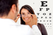Photo d'un opticien qu'on voit de dos tendant des lunettes pour une cliente souriante après avoir passer les tests de vues