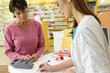 photo d'une patiente habillée en pull rouge et une pharmacienne donnant les médicaments à cette femme et notant le remboursement de la mutuelle santé sur papier