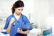 Photo d'une infirmière habillée en bleu tenant un sthetoscope et un dossier d'un patient qui se trouve derrière dans le lit d'hospitalisation